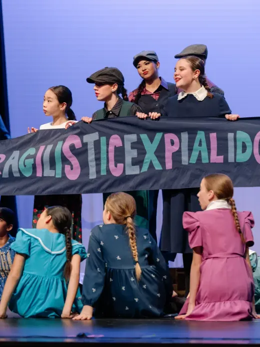 VPS Academy students performing Mary Poppins on stage holding a Supercalifragilisticexpialidocious banner